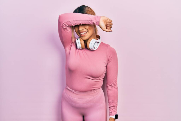 Beautiful hispanic woman wearing gym clothes and using headphones covering eyes with arm smiling cheerful and funny. blind concept.  - Photo, Image