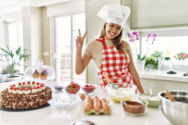 Güzel, genç, esmer pasta şefi kadın mutfakta hamur işleri pişirirken iki numarayı gösterip işaret ederken kendinden emin ve mutlu bir şekilde gülümsüyor..  - Fotoğraf, Görsel