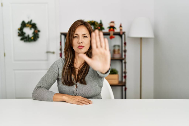 Jeune femme latine assise sur la table par décor de Noël faire arrêter chanter avec la paume de la main. expression d'avertissement avec geste négatif et sérieux sur le visage.  - Photo, image