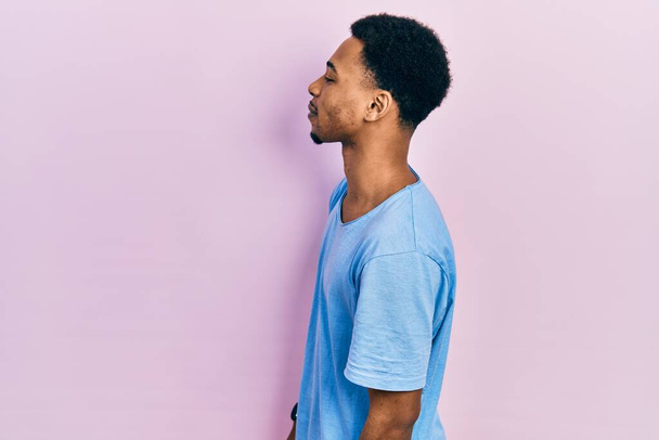 Afro-Amerikalı genç bir adam günlük mavi tişört giyiyor. Yan tarafa bakıyor, rahat bir poz veriyor ve kendine güvenen bir gülümseme takınıyor..  - Fotoğraf, Görsel