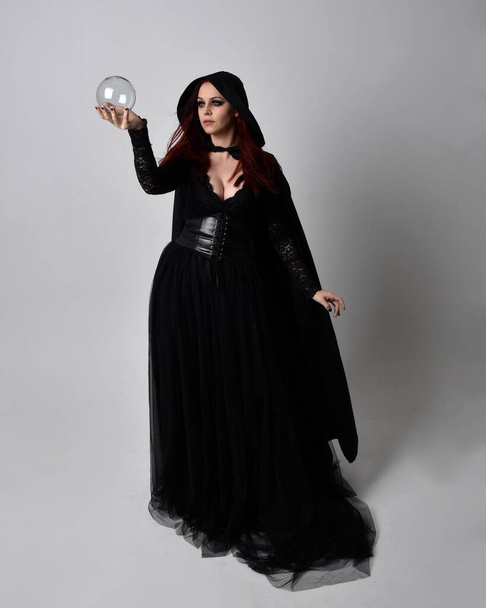 Volledig portret van een donkerharige vrouw in een zwart victoriaans heksenkostuum met een vloeiende mantel. staande houding, met gebaar handbewegingen, tegen studio achtergrond. - Foto, afbeelding