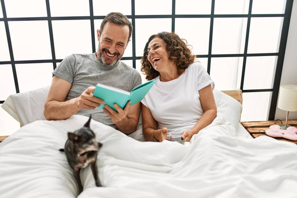 Middle age hispanic couple reading book and using smartphone. Lying on the bed with dog at home. - Photo, Image