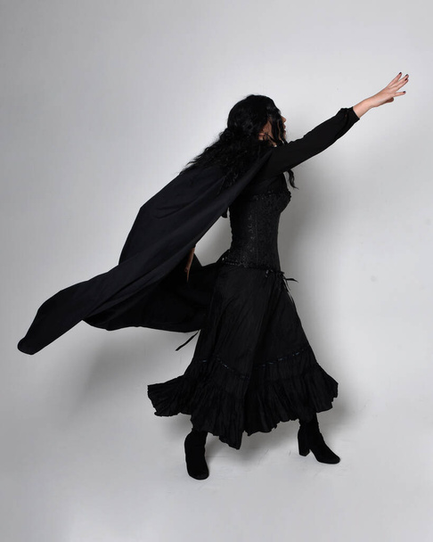 Full length portrait of dark haired woman wearing  black victorian witch costume with a flowing  cloak.  standing pose, with  gestural hand movements,  against studio background. - Foto, Imagem