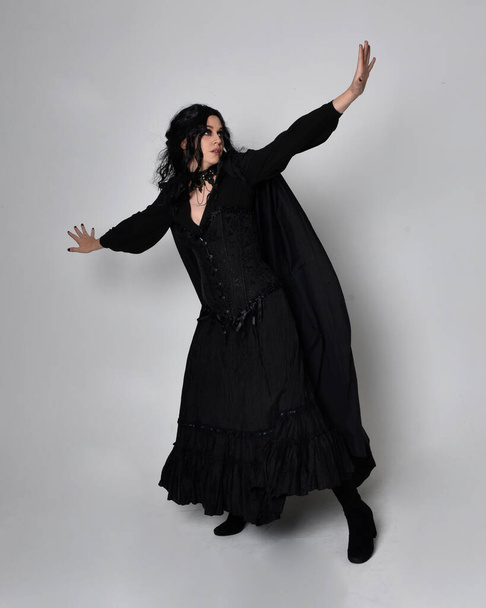 Full length portrait of dark haired woman wearing  black victorian witch costume with a flowing  cloak.  standing pose, with  gestural hand movements,  against studio background. - Foto, Bild