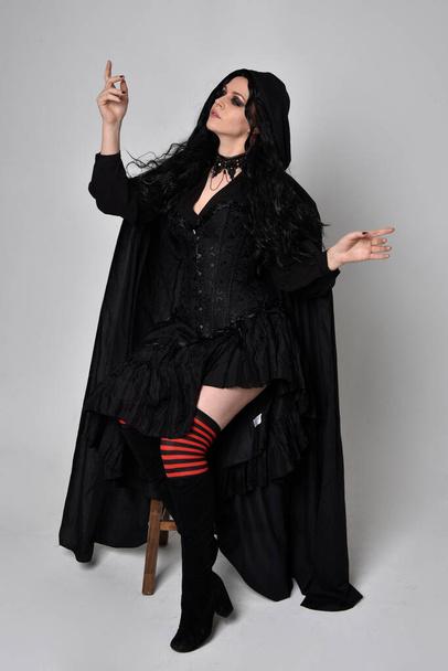 Full length portrait of dark haired woman wearing  black victorian witch costume with a flowing  cloak.  standing pose, with  gestural hand movements,  against studio background. - Foto, imagen