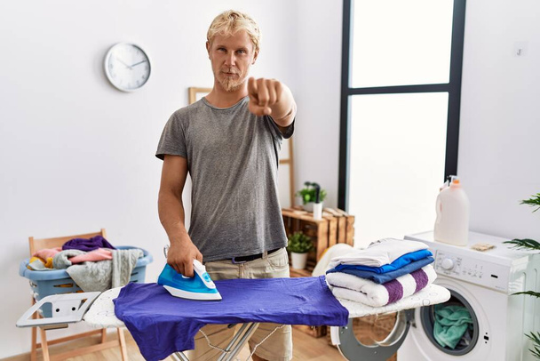 Young blond man ironing clothes at laundry room pointing with finger to the camera and to you, confident gesture looking serious  - Fotó, kép