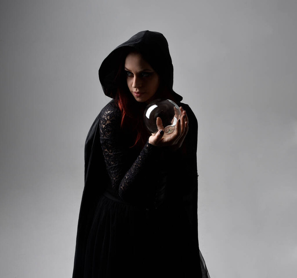 Siyah Victoria Cadı kostümü giyen kızıl saçlı kadının yakın plan portresi. Duruş, hareketli el hareketleri, karanlık stüdyo arka planı. - Fotoğraf, Görsel