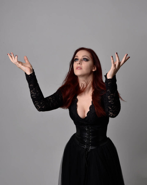 close up portrait of red haired woman wearing  black victorian witch costume.  standing pose, with  gestural hand movements,  against dark studio background. - Фото, изображение