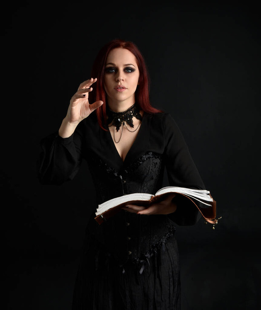 Siyah Victoria Cadı kostümü giyen kızıl saçlı kadının yakın plan portresi. Duruş, hareketli el hareketleri, karanlık stüdyo arka planı. - Fotoğraf, Görsel