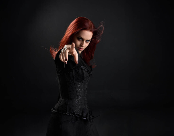 close up retrato de mulher de cabelos vermelhos vestindo traje de bruxa negra vitoriana. postura em pé, com movimentos gestuais das mãos, contra fundo escuro do estúdio. - Foto, Imagem