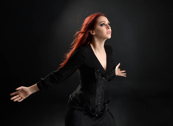 lähikuva punatukkainen nainen yllään musta Victorian noita puku. seisova asento, jossa eleellinen käsi liikkeet, vastaan tumma studio tausta. - Valokuva, kuva