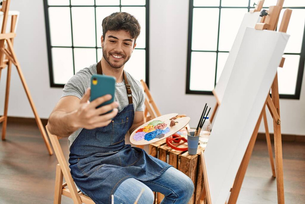 Junger hispanischer Künstler lächelt glücklich beim Malen und benutzt Smartphone im Kunstatelier. - Foto, Bild