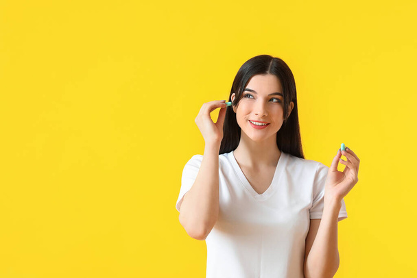 Pretty young woman putting blue ear plug on yellow background - Photo, Image