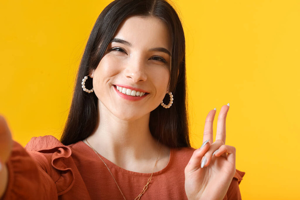 Hezká mladá žena přičemž selfie a ukazuje vítězství gesto na žlutém pozadí, detailní záběr - Fotografie, Obrázek