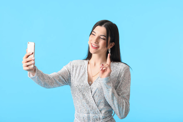 Kaunis nuori nainen ottaa selfie ja osoittaa voiton ele sinisellä taustalla - Valokuva, kuva