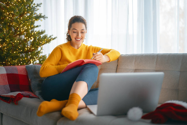Woman is making notes sitting on the sofa in the living room decorated for Christmas. - Foto, Bild