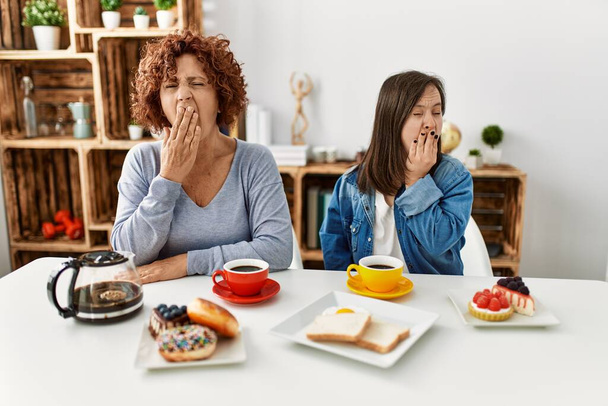 Rodzina matki i córki z zespołem Downa, siedzącej w domu jedzącej śniadanie znudzone ziewaniem zmęczone przykrywaniem ust dłonią. niepokój i senność.  - Zdjęcie, obraz