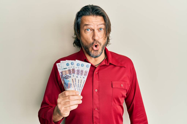 Pohledný muž středního věku drží ruské 500 rublů bankovky vyděšený a ohromený s otevřenou pusou pro překvapení, nevěřící tvář  - Fotografie, Obrázek