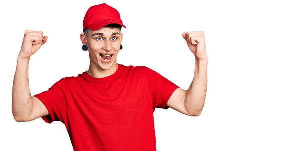 Giovane ragazzo caucasico con le orecchie dilatazione indossa uniforme consegna e berretto urlando orgoglioso, celebrando la vittoria e il successo molto eccitato con le braccia alzate  - Foto, immagini
