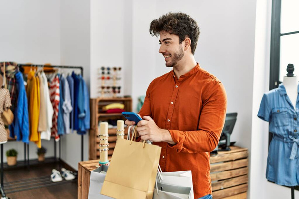 若いヒスパニック系の顧客の男笑顔幸せなショッピングバッグを保持し、衣料品店でスマートフォンを使用して. - 写真・画像