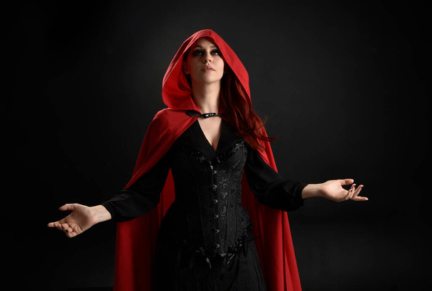 zbliżenie portret dziewczyny w czerwonym kapturze kaptur i strój czarownicy, izolowane na czarnym tle studio z zacienionym oświetleniem obręczy.  - Zdjęcie, obraz