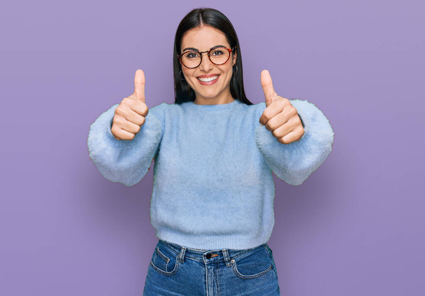 Mujer hispana joven usando ropa casual y gafas aprobando hacer gesto positivo con la mano, pulgares arriba sonriendo y feliz por el éxito. gesto ganador.  - Foto, imagen