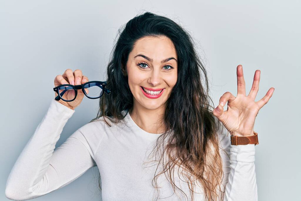 Молода іспаномовна жінка тримає окуляри роблячи знак з пальцями, посміхаючись дружній жест відмінний символ
  - Фото, зображення