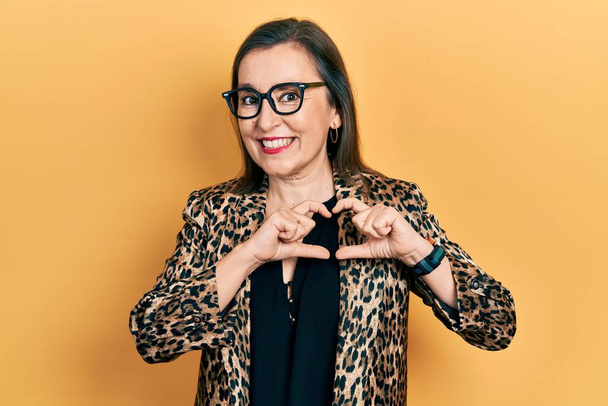 Középkorú spanyol nő visel üzleti ruhák és szemüveg mosolyog a szerelemben mutató szív szimbólum és alakja kézzel. romantikus koncepció.  - Fotó, kép