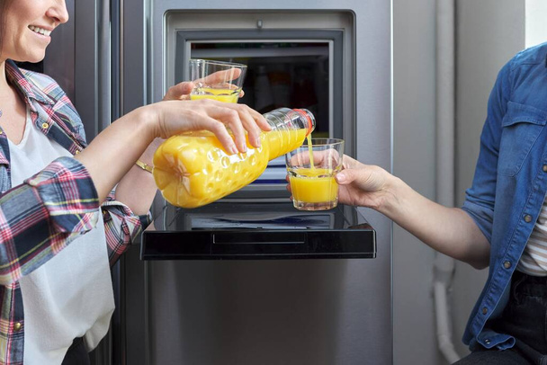 Gros plan de deux mains de femmes versant du jus d'orange dans le verre du réfrigérateur dans la cuisine - Photo, image