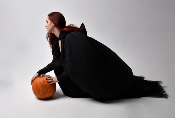 Plně dlouhý portrét tmavovlasé ženy v černém viktoriánském kostýmu sedícího čarodějnice, s gesturálními pohyby rukou, na pozadí ateliéru. - Fotografie, Obrázek