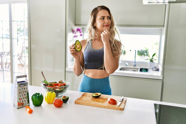 Kafkasyalı spor kıyafetleri giyen genç kadın mutfakta sağlıklı salata hazırlıyor ciddi bir yüz ifadesi takınıyor eli çenesinde soruyu düşünüyor, kafa karıştırıcı bir fikir.  - Fotoğraf, Görsel