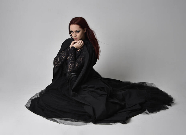 Ganztägiges Porträt einer dunkelhaarigen Frau im schwarzen Hexenkostüm sitzend, mit gestischen Handbewegungen, vor Studiohintergrund. - Foto, Bild