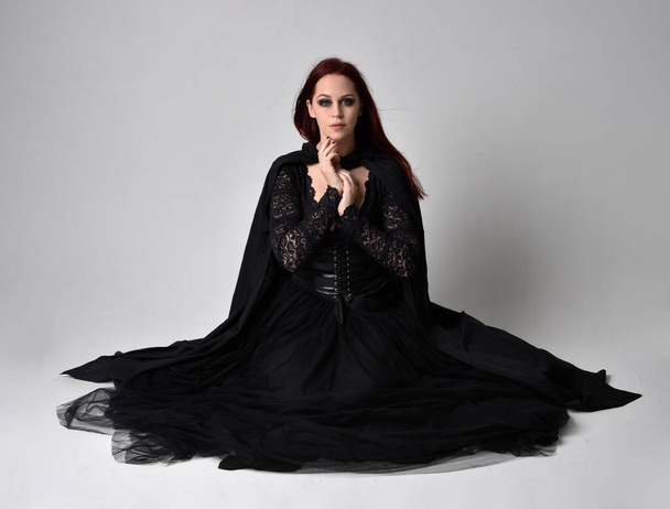 Ganztägiges Porträt einer dunkelhaarigen Frau im schwarzen Hexenkostüm sitzend, mit gestischen Handbewegungen, vor Studiohintergrund. - Foto, Bild