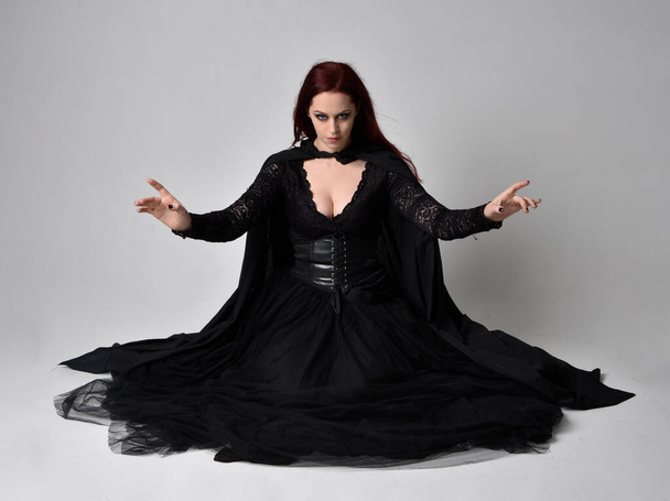 Pełnowymiarowy portret ciemnowłosej kobiety w czarnym wiktoriańskim kostiumie czarownicy siedzącej, z gestykulacyjnymi ruchami dłoni, na tle studia. - Zdjęcie, obraz