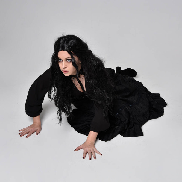 Полноразмерный портрет темноволосой женщины в черном костюме ведьмы, сидящей на студийном фоне, с жестикулирующими движениями рук. - Фото, изображение