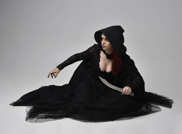 Retrato de comprimento total de mulher de cabelos escuros vestindo traje de bruxa preto vitoriano sentado pose, com movimentos gestuais das mãos, contra o fundo do estúdio. - Foto, Imagem