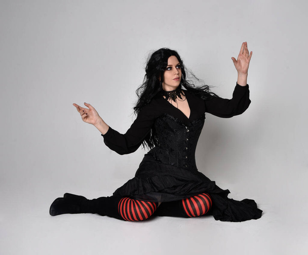 Полноразмерный портрет темноволосой женщины в черном костюме ведьмы, сидящей на студийном фоне, с жестикулирующими движениями рук. - Фото, изображение
