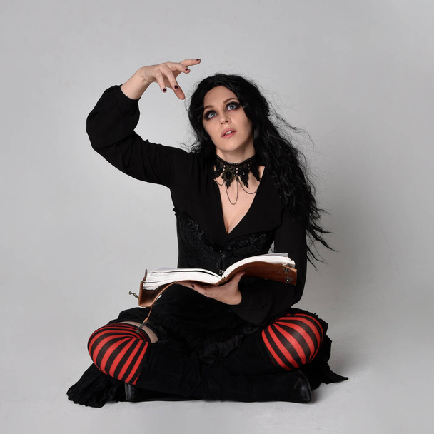Pełnowymiarowy portret ciemnowłosej kobiety w czarnym wiktoriańskim kostiumie czarownicy siedzącej, z gestykulacyjnymi ruchami dłoni, na tle studia. - Zdjęcie, obraz