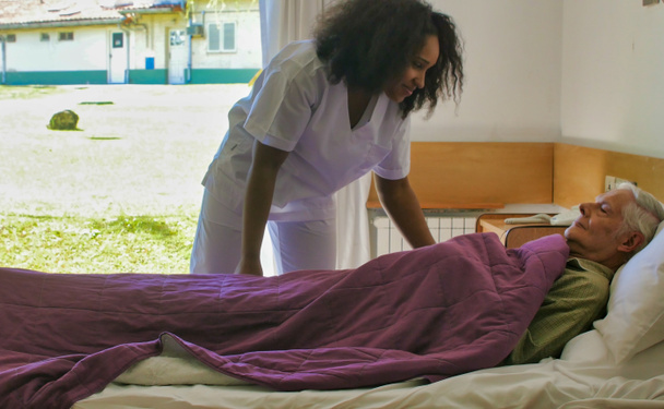 Genç Afrikalı kadın doktor hastane yatağında yaşlı bir adama yardım ediyor.. - Fotoğraf, Görsel