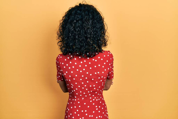 Joven chica latina con vestido de verano de pie hacia atrás mirando hacia otro lado con los brazos cruzados  - Foto, imagen