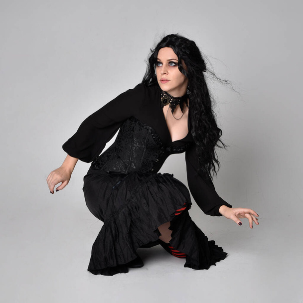 Full length portrait of dark haired woman wearing  black victorian witch costume  sitting pose, with  gestural hand movements,  against studio background. - Фото, зображення