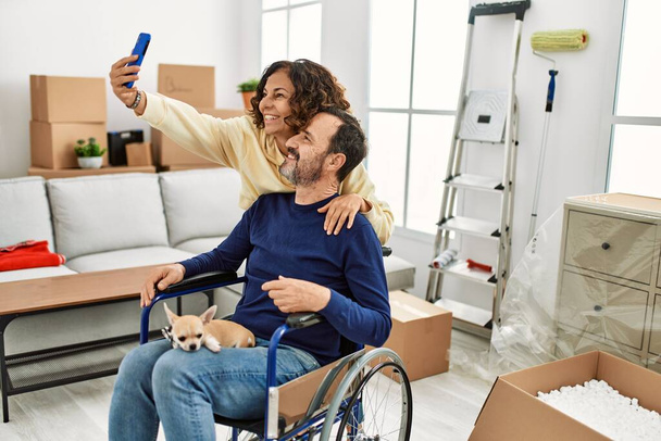 Orta yaşlı İspanyol çift mutlu gülümsüyor. Tekerlekli sandalyede oturan bir adam, bacaklarında köpek ve yeni evinde akıllı telefondan selfie çeken bir kadın.. - Fotoğraf, Görsel
