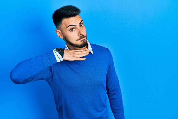 Jonge Spaanse man met baard die casual blauwe trui draagt en keel doorsnijdt als mes, bedreigt agressie met woedend geweld.  - Foto, afbeelding