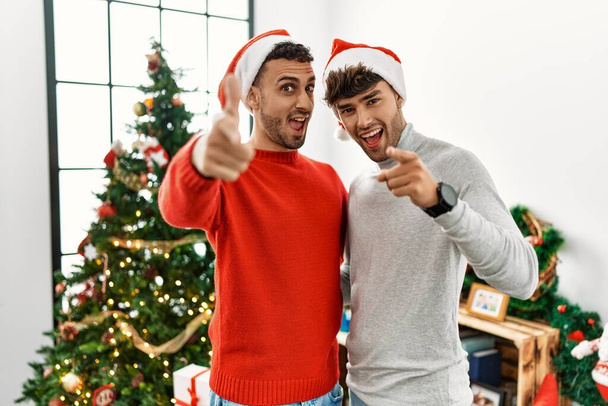 Young gay couple standing by christmas tree wearing hat pointing fingers to camera with happy and funny face. good energy and vibes.  - Photo, Image
