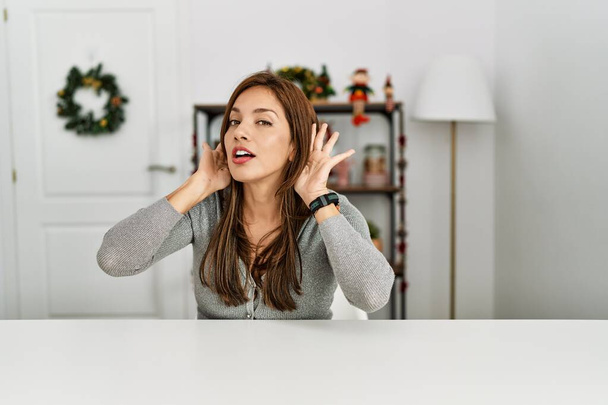 Genç Latin kadın Noel dekoru yüzünden masada oturmuş iki elini de dedikodu meraklısı bir şekilde kulak hareketi yaparken duymaya çalışıyor. İşitme sorunu, sağır  - Fotoğraf, Görsel