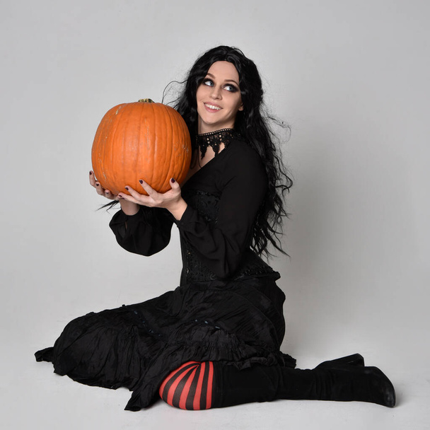 Pełnowymiarowy portret ciemnowłosej kobiety ubranej w czarny wiktoriański kostium czarownicy siedzącej pozy wit ha dyni, z gestywnymi ruchami dłoni, na tle studia. - Zdjęcie, obraz