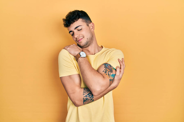 Jonge Latijns-Amerikaanse man draagt casual t-shirt knuffelend zichzelf gelukkig en positief, glimlachend zelfverzekerd. zelfliefde en zelfzorg  - Foto, afbeelding