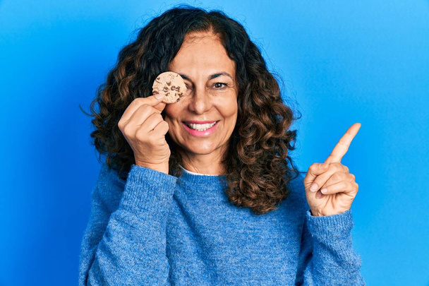 Латиноамериканка средних лет держит печенье над глазом улыбаясь счастливо указывая рукой и пальцем в сторону  - Фото, изображение