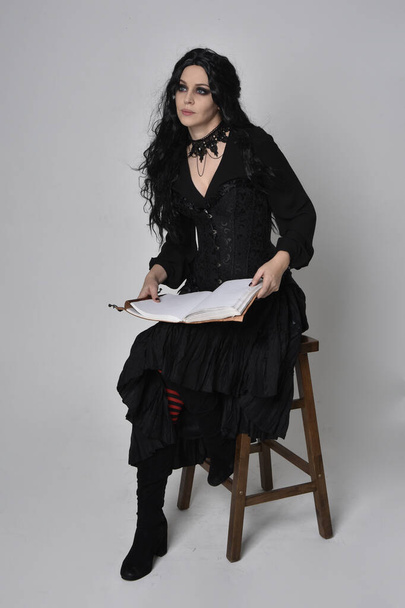 Retrato de cuerpo entero de mujer morena vestida con traje de bruja victoriana negra. posar sentado en una silla, con movimientos gestuales de la mano, contra el fondo del estudio. - Foto, Imagen