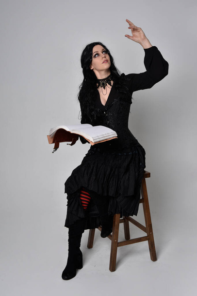 Siyah Victoria cadısı kostümü giymiş koyu renk saçlı bir kadının tam boy portresi. Sandalyede otururken, el hareketleriyle, stüdyo arka planında.. - Fotoğraf, Görsel